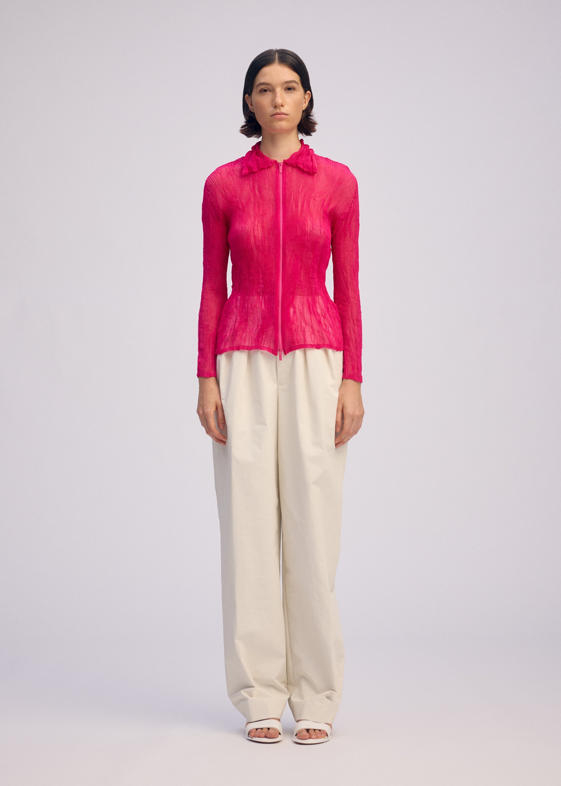 Zoomed view A woman with short, dark hair faces forward against a plain background, wearing the ISSEY MIYAKE CHIFFON TWIST 1 bright pink pleated zip-up cardigan with a collar, paired with loose high-waisted cream trousers and white open-toe shoes.