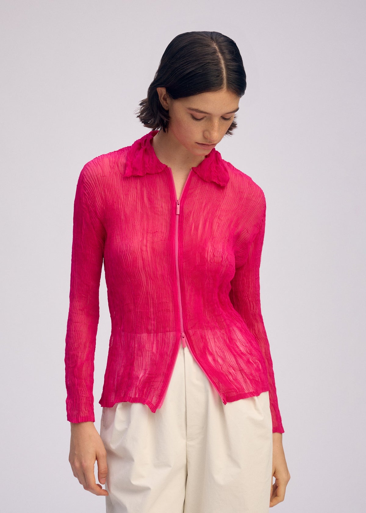 A person with short dark hair wears the ISSEY MIYAKE CHIFFON TWIST 1, a bright pink pleated zip-up cardigan, paired with high-waisted white pants. They stand against a plain light background, looking downward. view 6 of 7
