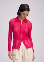 A person with short dark hair wears the ISSEY MIYAKE CHIFFON TWIST 1, a bright pink pleated zip-up cardigan, paired with high-waisted white pants. They stand against a plain light background, looking downward.