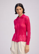 A woman with short dark hair wears ISSEY MIYAKE’s CHIFFON TWIST 1, a sheer bright pink zip-up cardigan, with loose white pants against a plain light gray background.