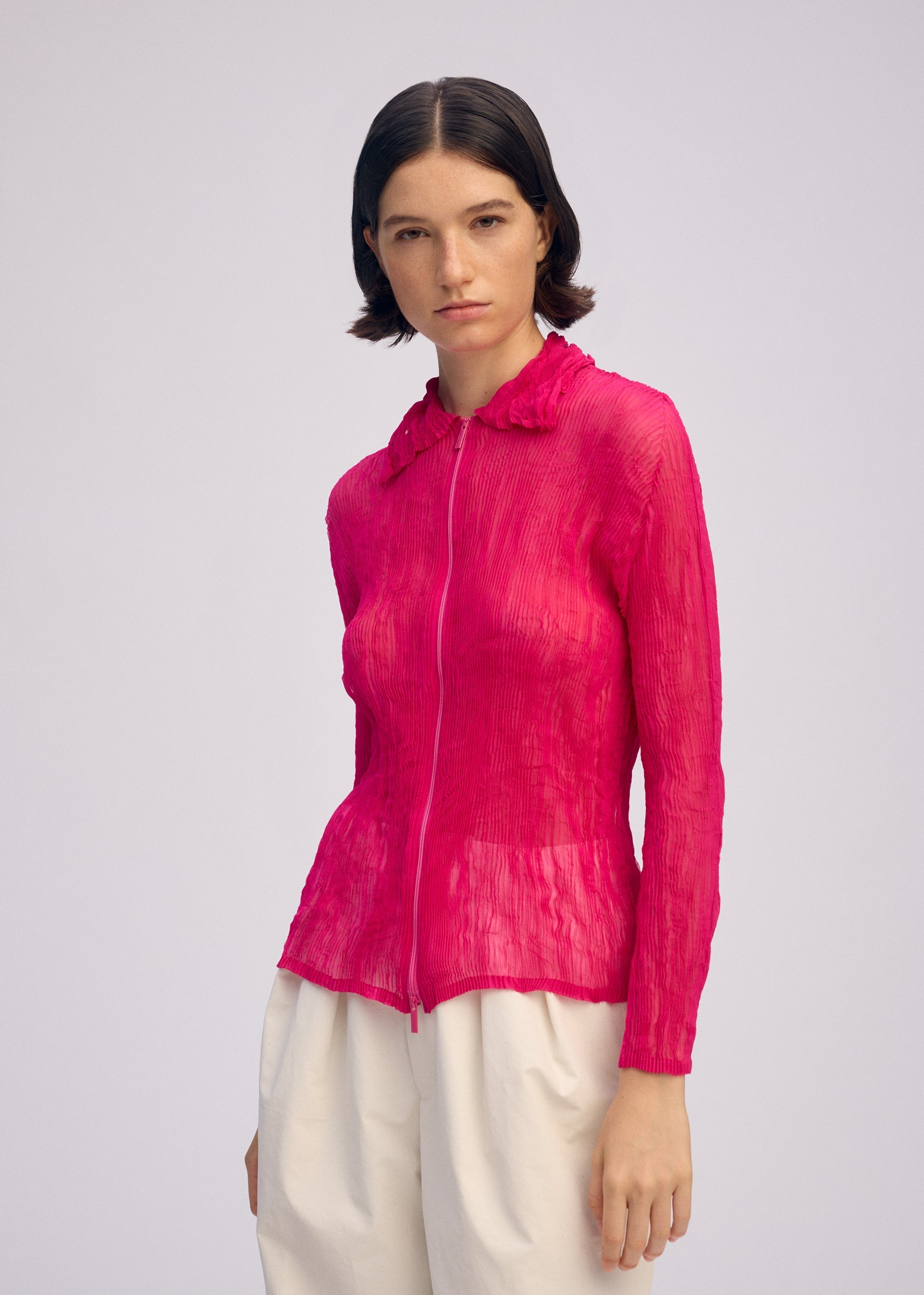 Zoomed view A woman with short dark hair wears ISSEY MIYAKE’s CHIFFON TWIST 1, a sheer bright pink zip-up cardigan, with loose white pants against a plain light gray background.