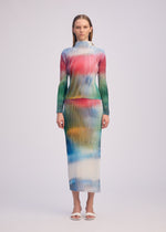 A woman wears ISSEY MIYAKE’s OBSCURE PLEATS: a long-sleeved, high-neck pleated polyester top and ankle-length dress with a vibrant frosted glass print in red, green, blue, and yellow, paired with white open-toe sandals.