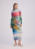 A woman wears the OBSCURE PLEATS dress by ISSEY MIYAKE, a long-sleeved, high-neck ribbed piece with a pink, blue, green, yellow, and white gradient evoking frosted glass. She pairs it with white heeled sandals against a plain background.