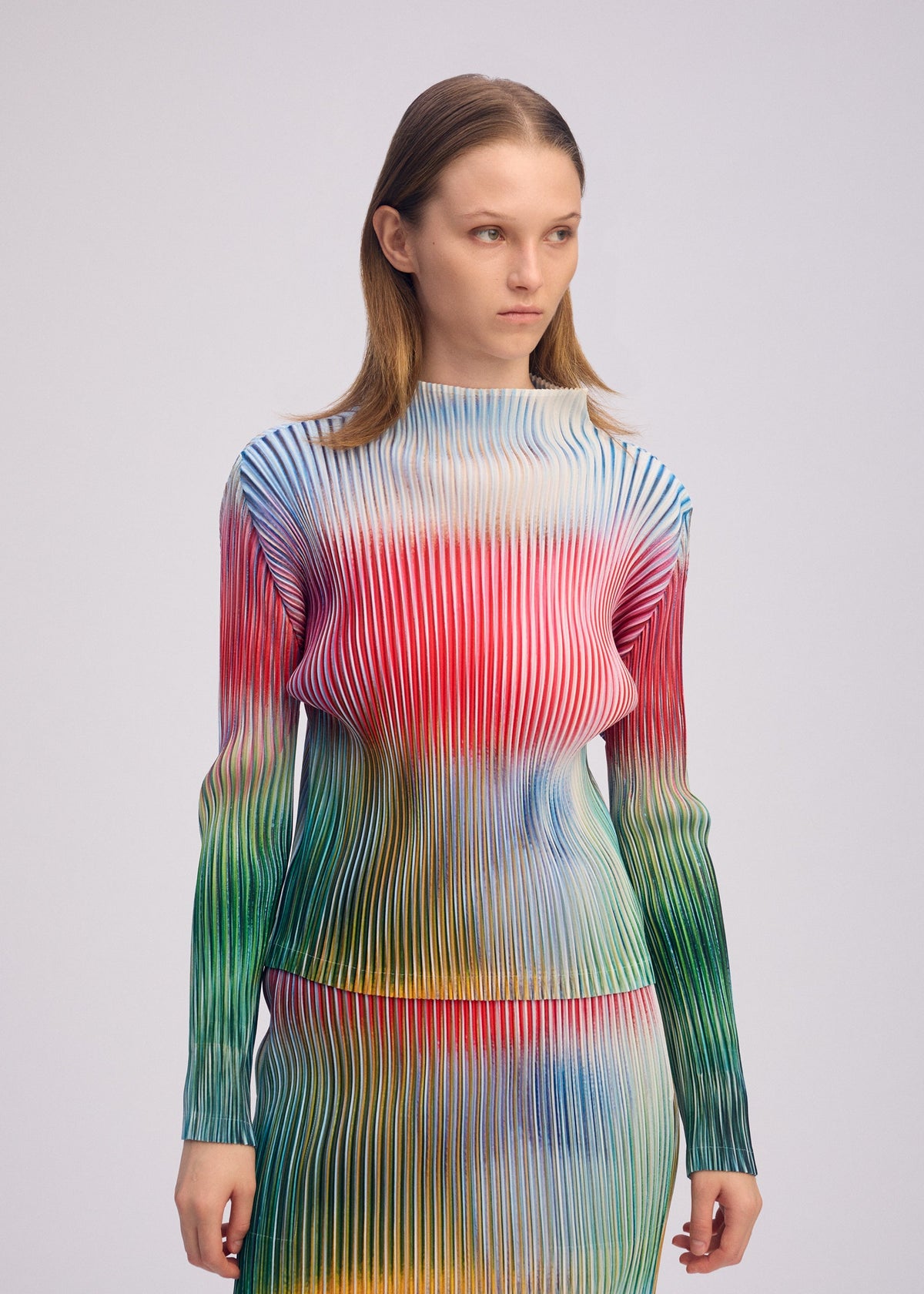 A person wears the OBSCURE PLEATS top and skirt by ISSEY MIYAKE, featuring long sleeves, a high neck, and colorful vertical wavy moiré stripes in red, blue, green, and yellow, standing against a plain light background. view 8 of 8