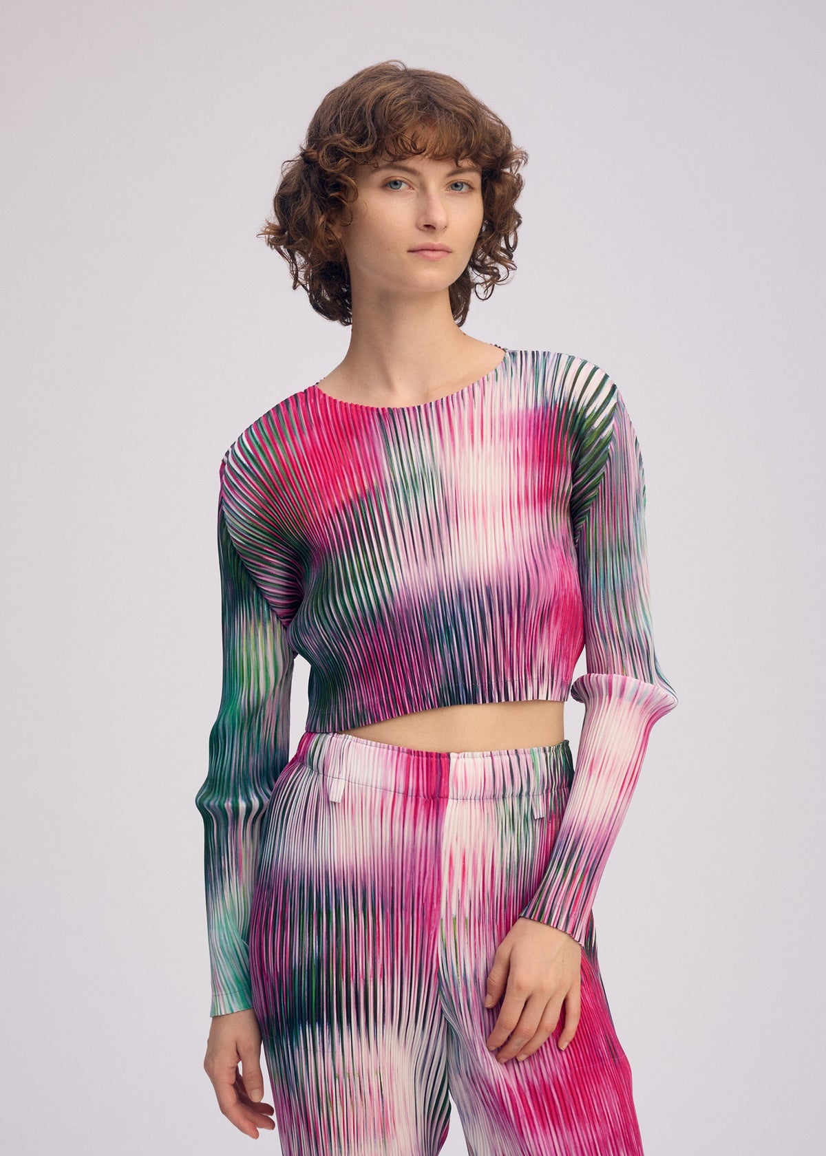 A person with short curly hair wears the ISSEY MIYAKE OBSCURE PLEATS set—pleated polyester long-sleeved top and pants in a vibrant pink, green, and white gradient—against a plain light background. view 7 of 7