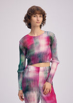 A person with short curly hair wears the ISSEY MIYAKE OBSCURE PLEATS set—pleated polyester long-sleeved top and pants in a vibrant pink, green, and white gradient—against a plain light background.