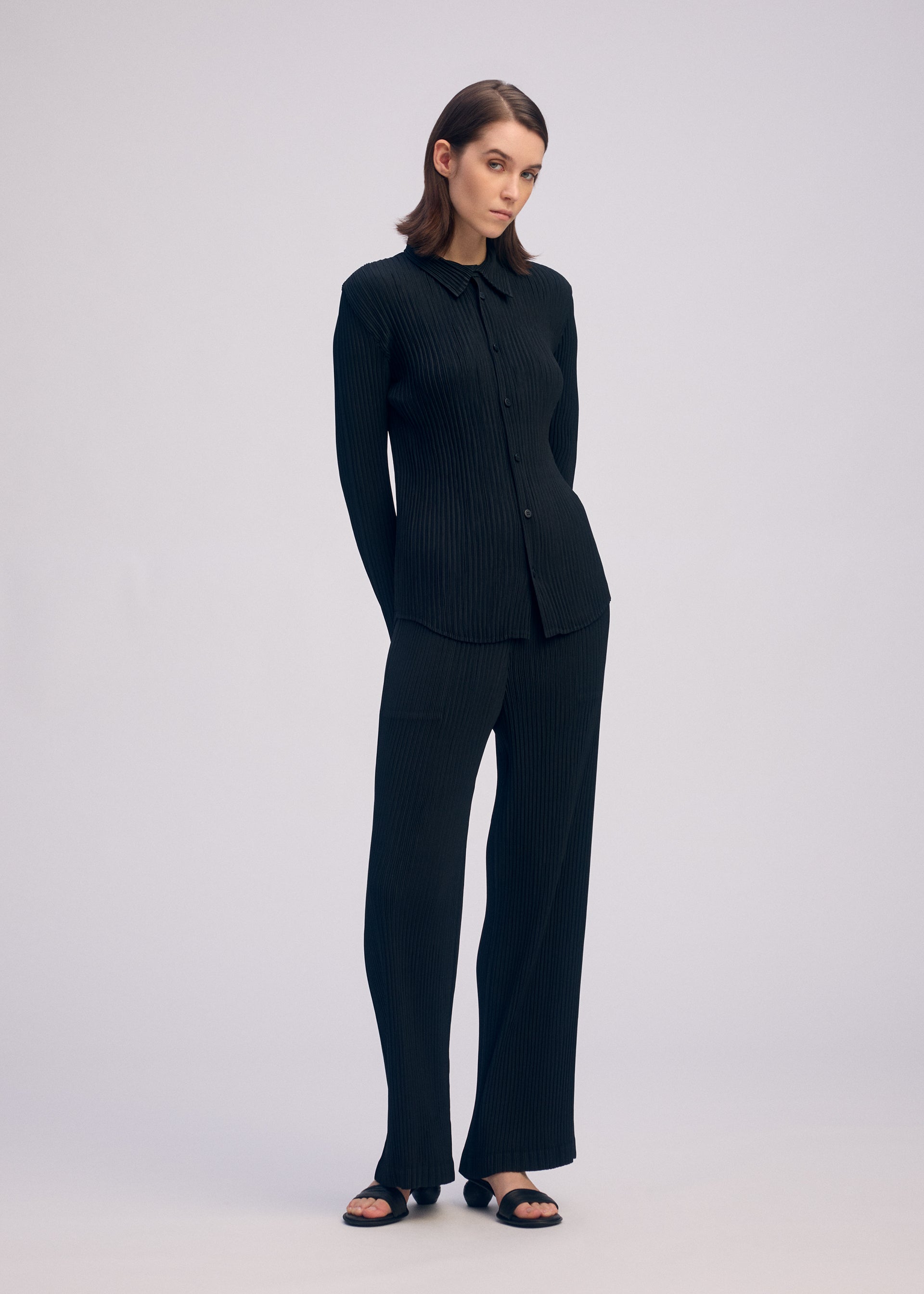 Zoomed view A woman stands against a plain light background, wearing the ISSEY MIYAKE HATCHING PLEATS black long-sleeve collared shirt and matching wide-leg trousers with a subtle ribbed texture, paired with black sandals. Her hands are behind her back.