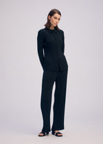 A woman with short brown hair stands against a plain background in ISSEY MIYAKE’s HATCHING PLEATS set: a black long-sleeve recycled polyester shirt and matching textured wide-leg pants, styled with black open-toe sandals—ideal for daily wear.