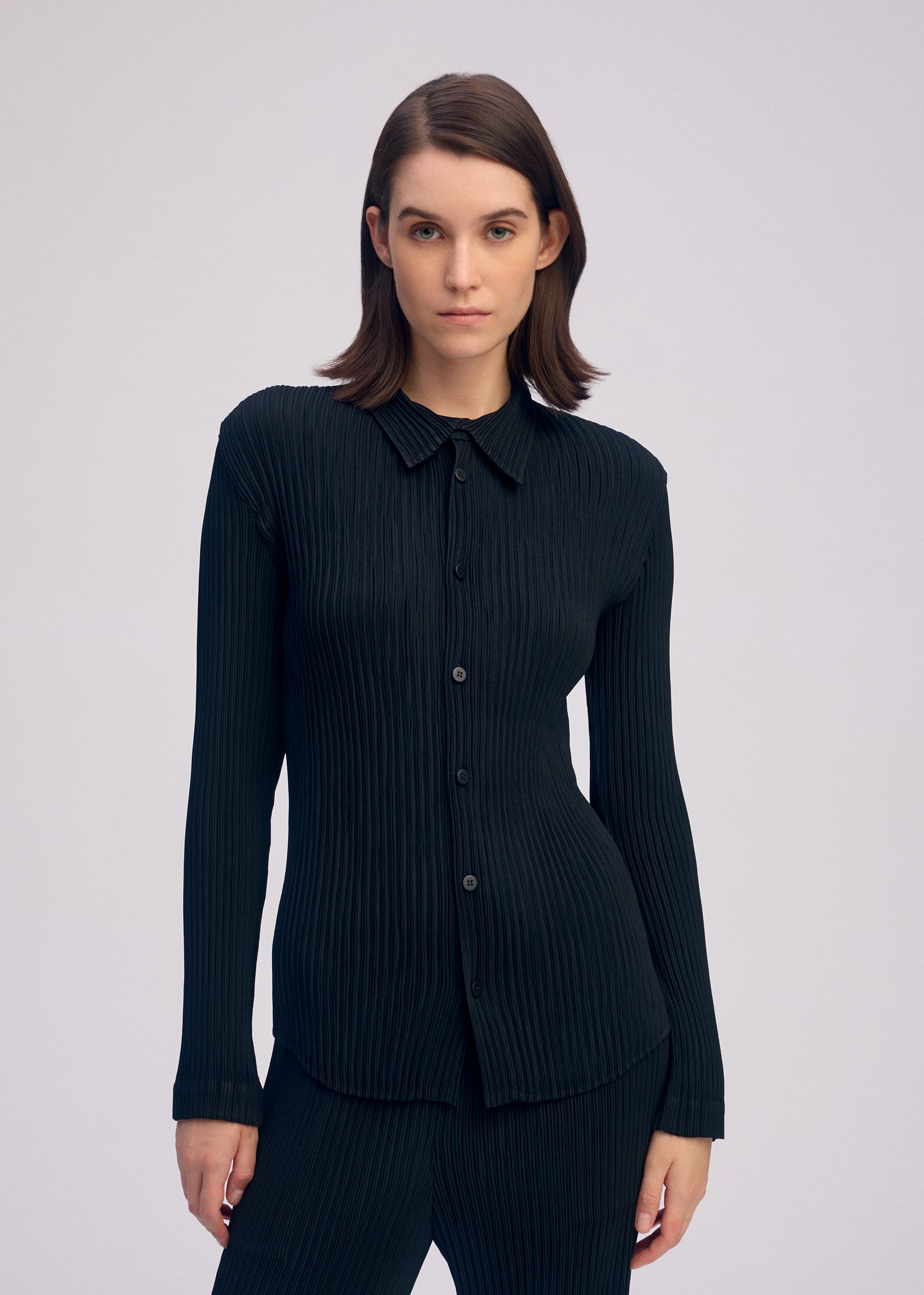 Zoomed view A person with straight brown hair stands facing forward against a plain light background, wearing ISSEY MIYAKE's HATCHING PLEATS: a black, long-sleeved pleated shirt and matching textured pants.