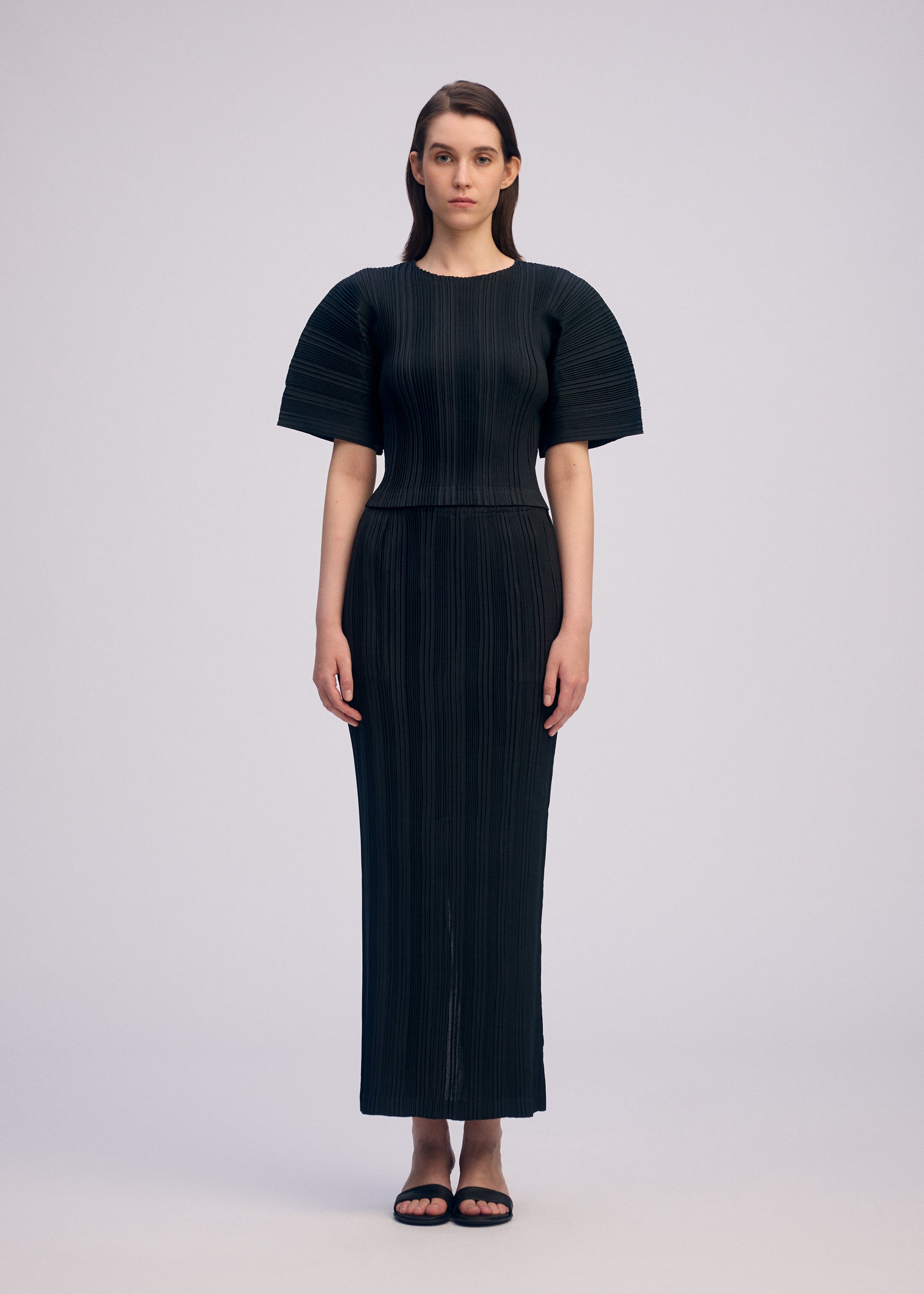 Zoomed view A woman with straight brown hair wears ISSEY MIYAKE's LINEN LIKE RIB PLEATS: a fitted, round-neck pleated top with structured shoulders and a matching long skirt. She pairs the look with black sandals against a plain, light background.