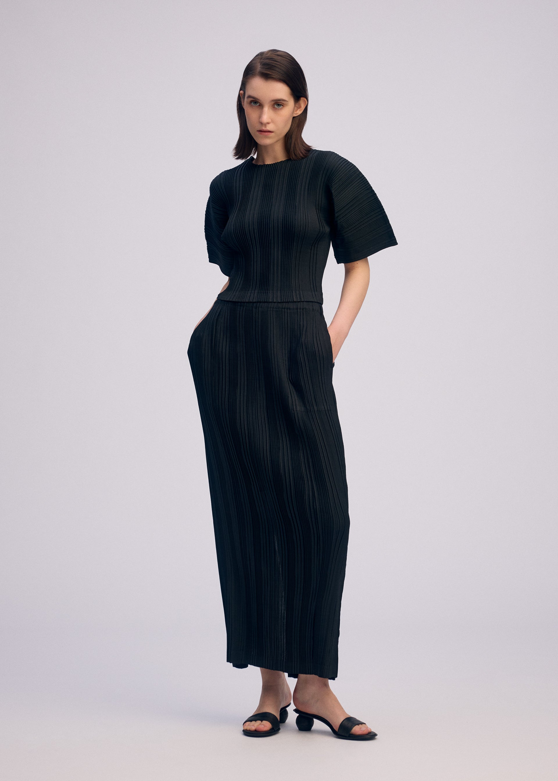 Zoomed view A woman stands against a plain background wearing the ISSEY MIYAKE LINEN LIKE RIB PLEATS ankle-length, short-sleeved black dress with subtle vertical stripes. She has shoulder-length straight hair, black sandals, and her hands in her pockets.