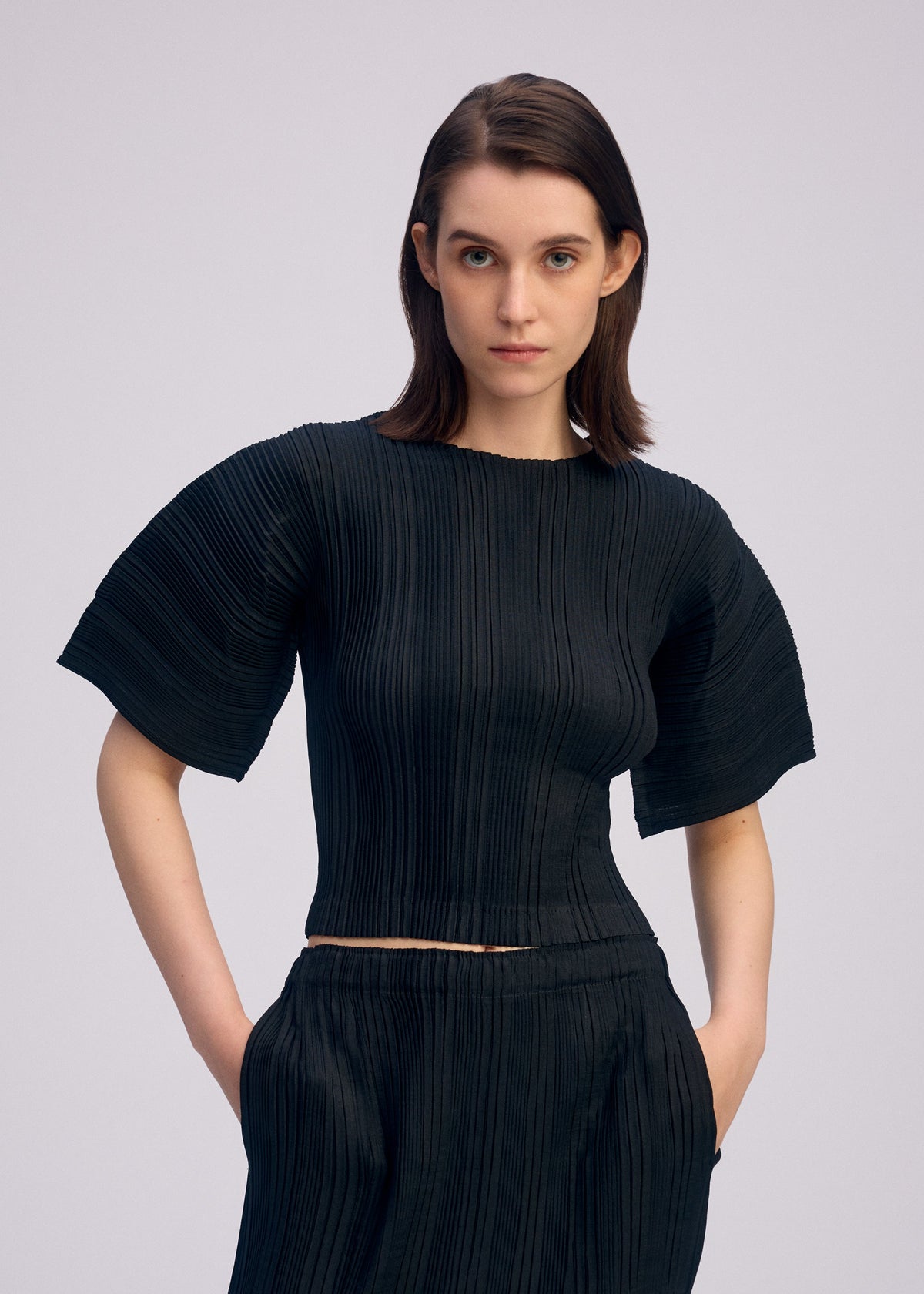 A woman with straight brown hair models the ISSEY MIYAKE LINEN LIKE RIB PLEATS black round-neck pleated top and matching pleated pants, standing with hands in her pockets against a plain light backdrop. view 7 of 8