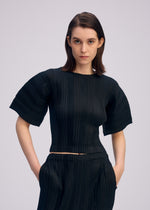 A woman with straight brown hair models the ISSEY MIYAKE LINEN LIKE RIB PLEATS black round-neck pleated top and matching pleated pants, standing with hands in her pockets against a plain light backdrop.