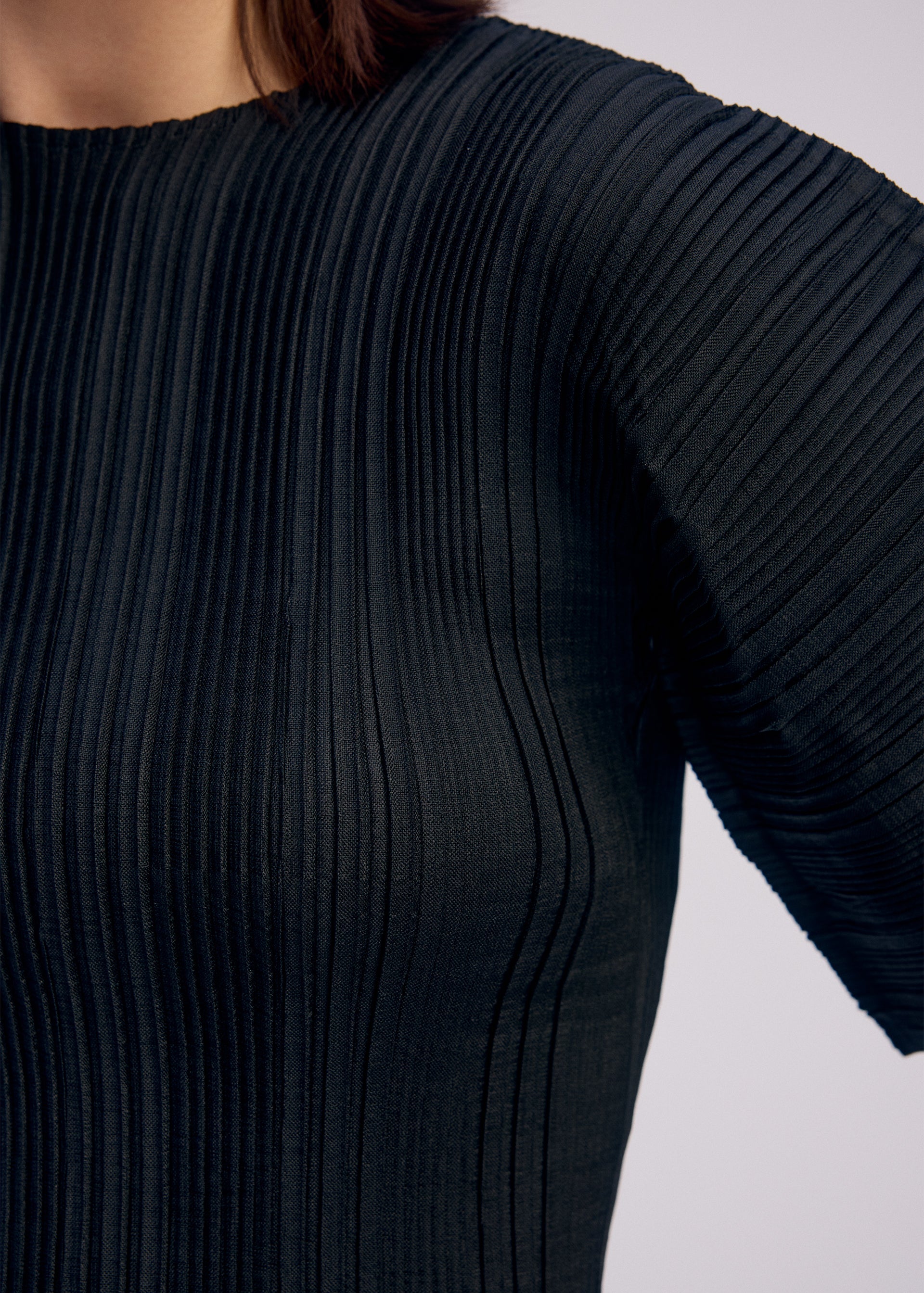 Zoomed view Close-up of a person wearing the ISSEY MIYAKE LINEN LIKE RIB PLEATS top, featuring black, pleated fabric with vertical ribbed texture. The image shows the shoulder, upper chest, and part of the arm in this stylish short-sleeve blouse.
