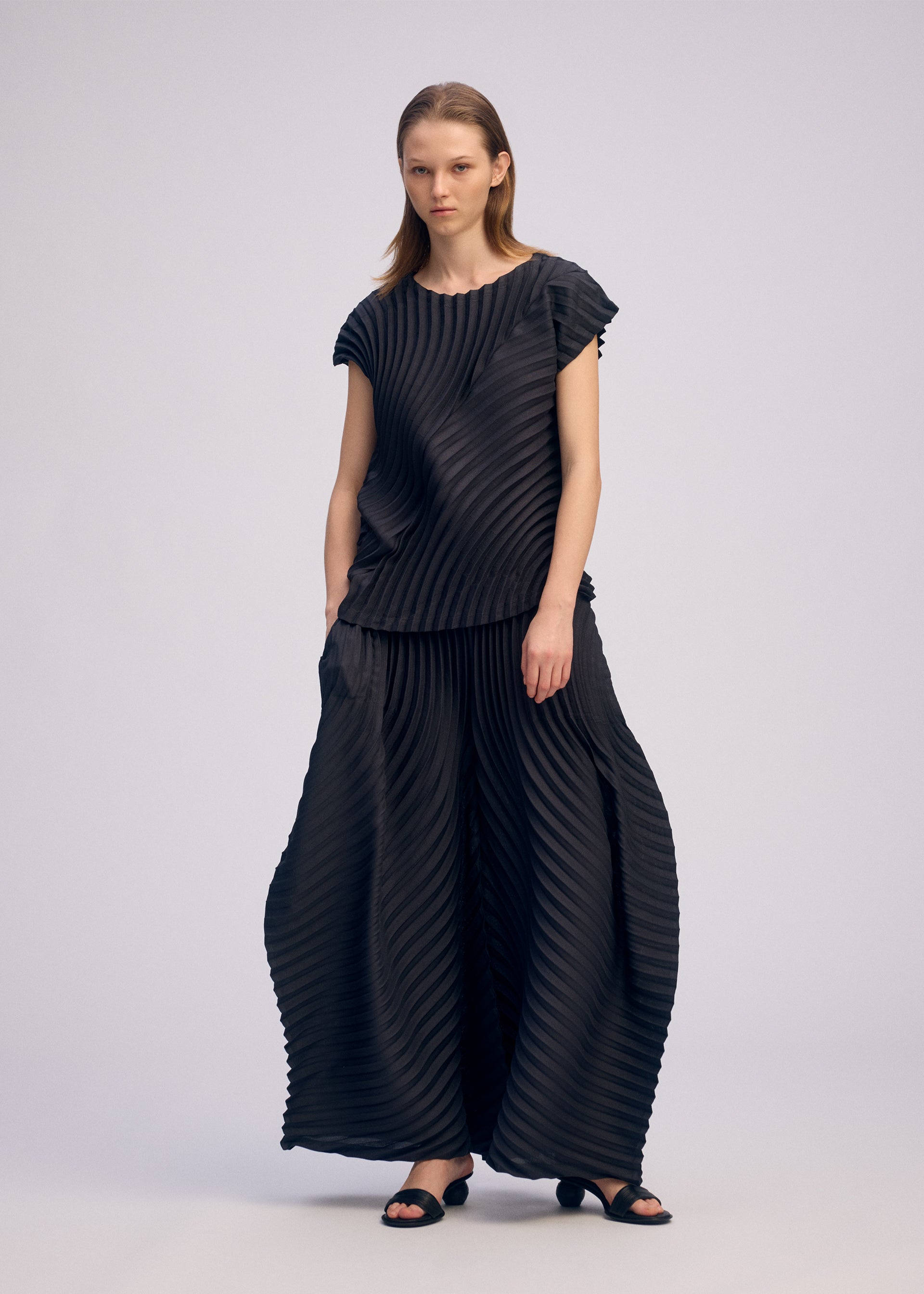 Zoomed view A woman with straight, shoulder-length hair wears ISSEY MIYAKE's CIRCLE PLEATS PB: a round-neck black sleeveless top and wide-legged pants with handmade pleats, paired with classic black sandals against a plain background.