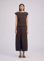 A woman faces forward in the ISSEY MIYAKE PLASTER PLEATS SOLID set, featuring a brown box-pleat short-sleeve top and wide-leg pants. She wears black sandals, has straight dark hair, and stands against a plain light background.