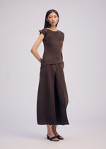A woman stands against a plain background, wearing ISSEY MIYAKE's PLASTER PLEATS SOLID set—a matching dark brown crêpe de Chine top and wide-leg pants. She has straight dark hair, hands behind her back, and wears black open-toe sandals.