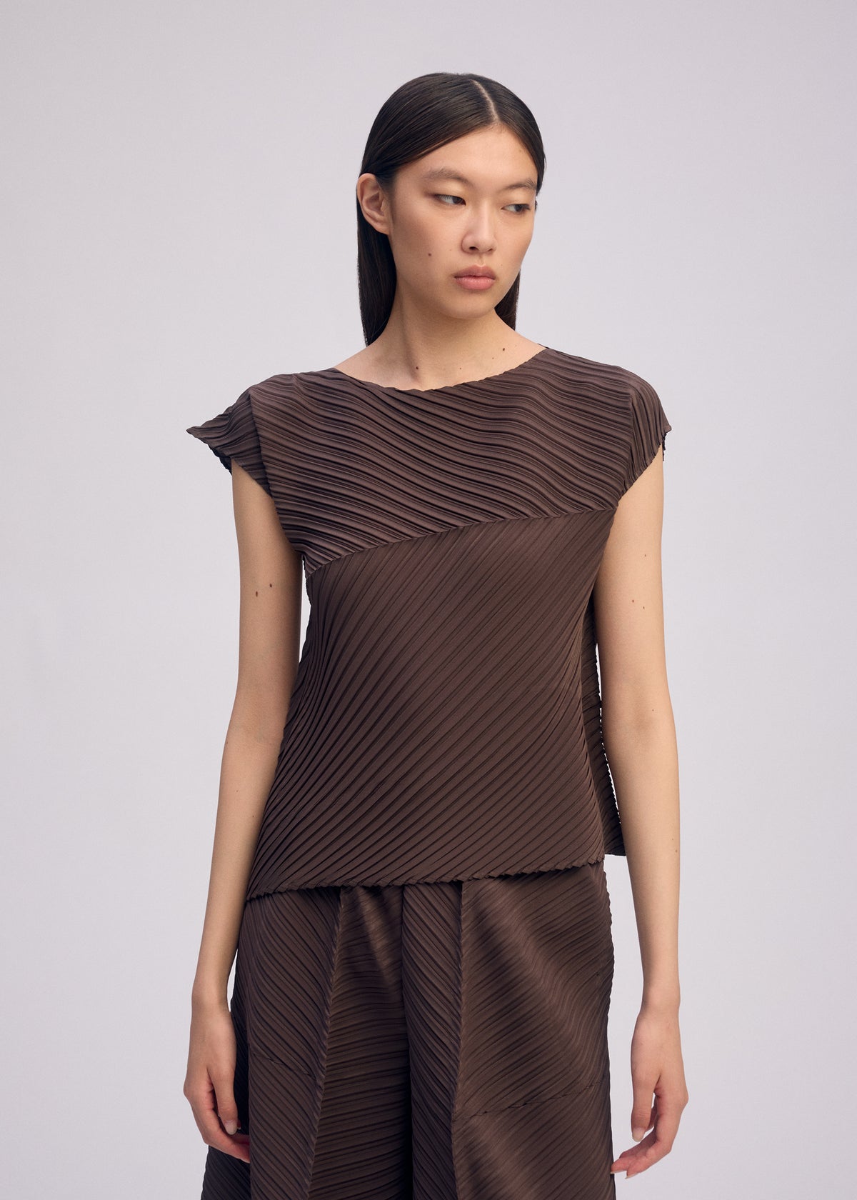 A woman with straight dark hair wears the ISSEY MIYAKE PLASTER PLEATS SOLID set—brown box pleat crêpe de Chine top and matching wide-leg pants—standing against a plain light background, gazing slightly to the side with a neutral expression. view 7 of 7