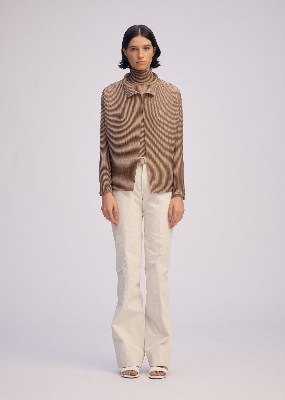 A person stands facing forward in the WOOLY PLEATS-66 brown pleated cardigan by ISSEY MIYAKE, over a brown turtleneck with white high-waisted belted trousers and white open-toe heels, against a plain light background. view 5 of 7