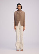 A person stands facing forward in the WOOLY PLEATS-66 brown pleated cardigan by ISSEY MIYAKE, over a brown turtleneck with white high-waisted belted trousers and white open-toe heels, against a plain light background.