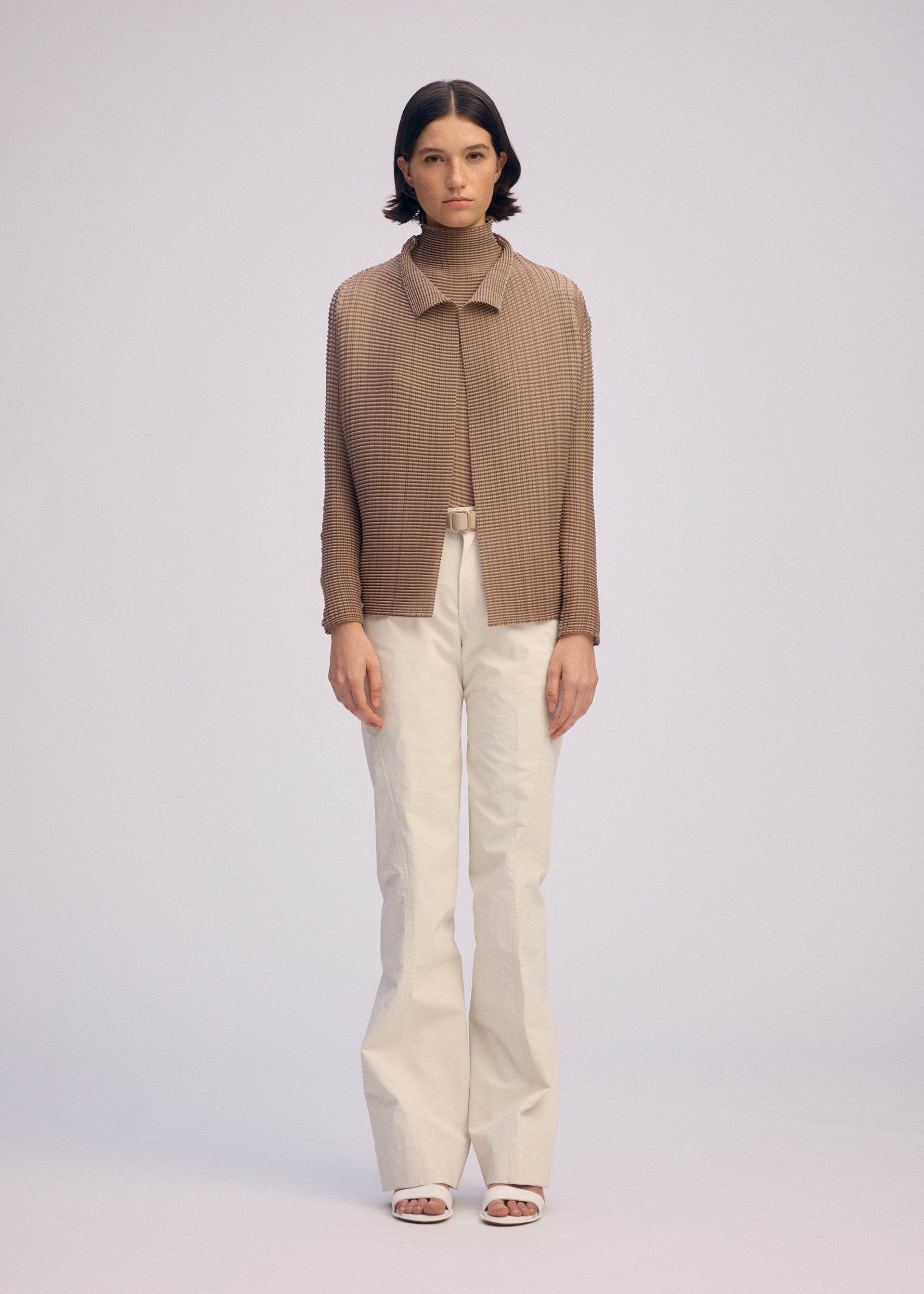 Zoomed view A person stands facing forward in the WOOLY PLEATS-66 brown pleated cardigan by ISSEY MIYAKE, over a brown turtleneck with white high-waisted belted trousers and white open-toe heels, against a plain light background.