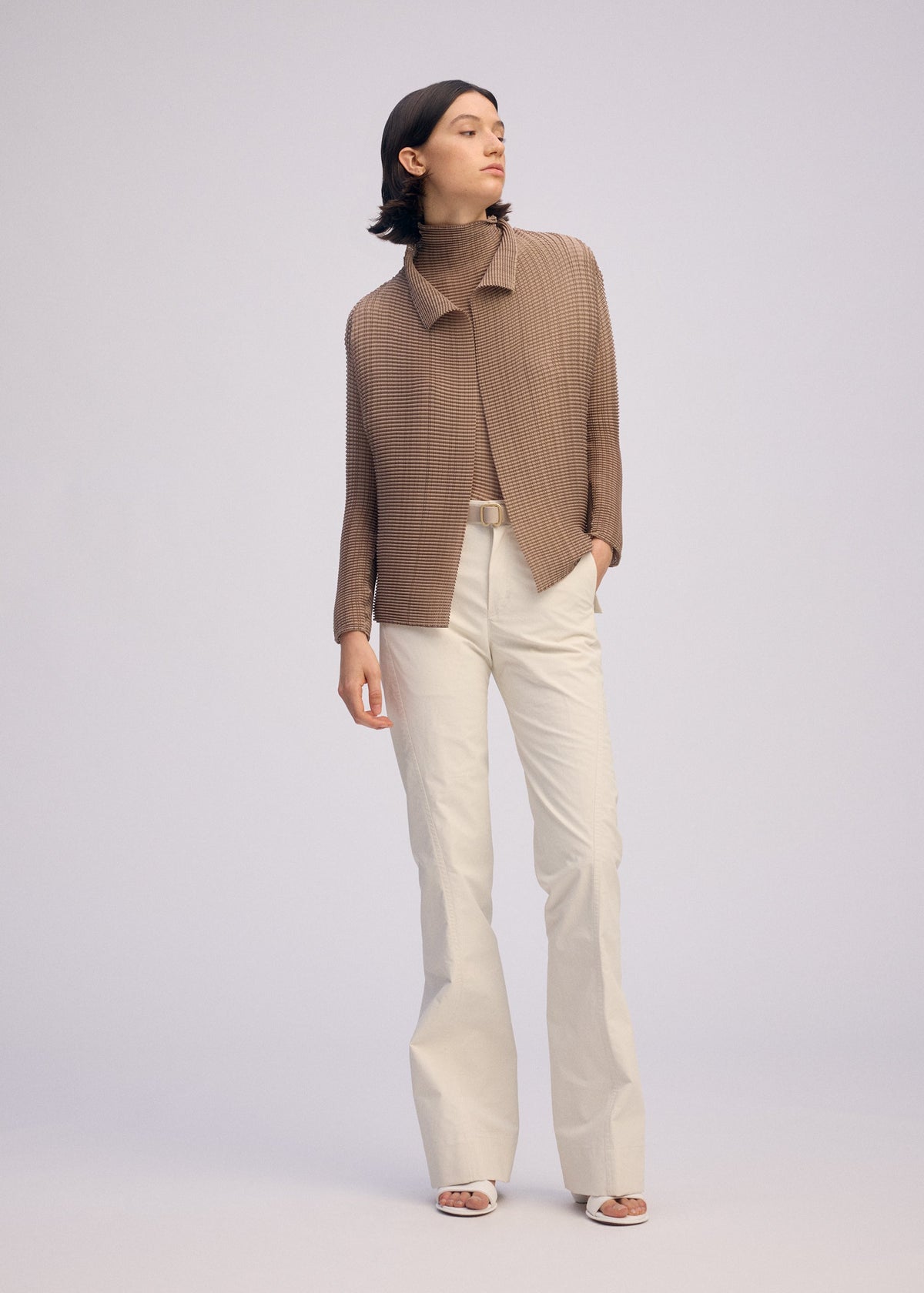 A woman with short dark hair stands against a plain background, wearing the ISSEY MIYAKE WOOLY PLEATS-66 brown pleated cardigan over a matching turtleneck, paired with cream pants and white sandals, looking to the side with one hand in her pocket. view 6 of 7