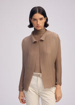A woman with short dark hair wears the ISSEY MIYAKE WOOLY PLEATS-66, a textured light brown pleated cardigan over a matching turtleneck and cream pants with a belt, standing against a plain light background.