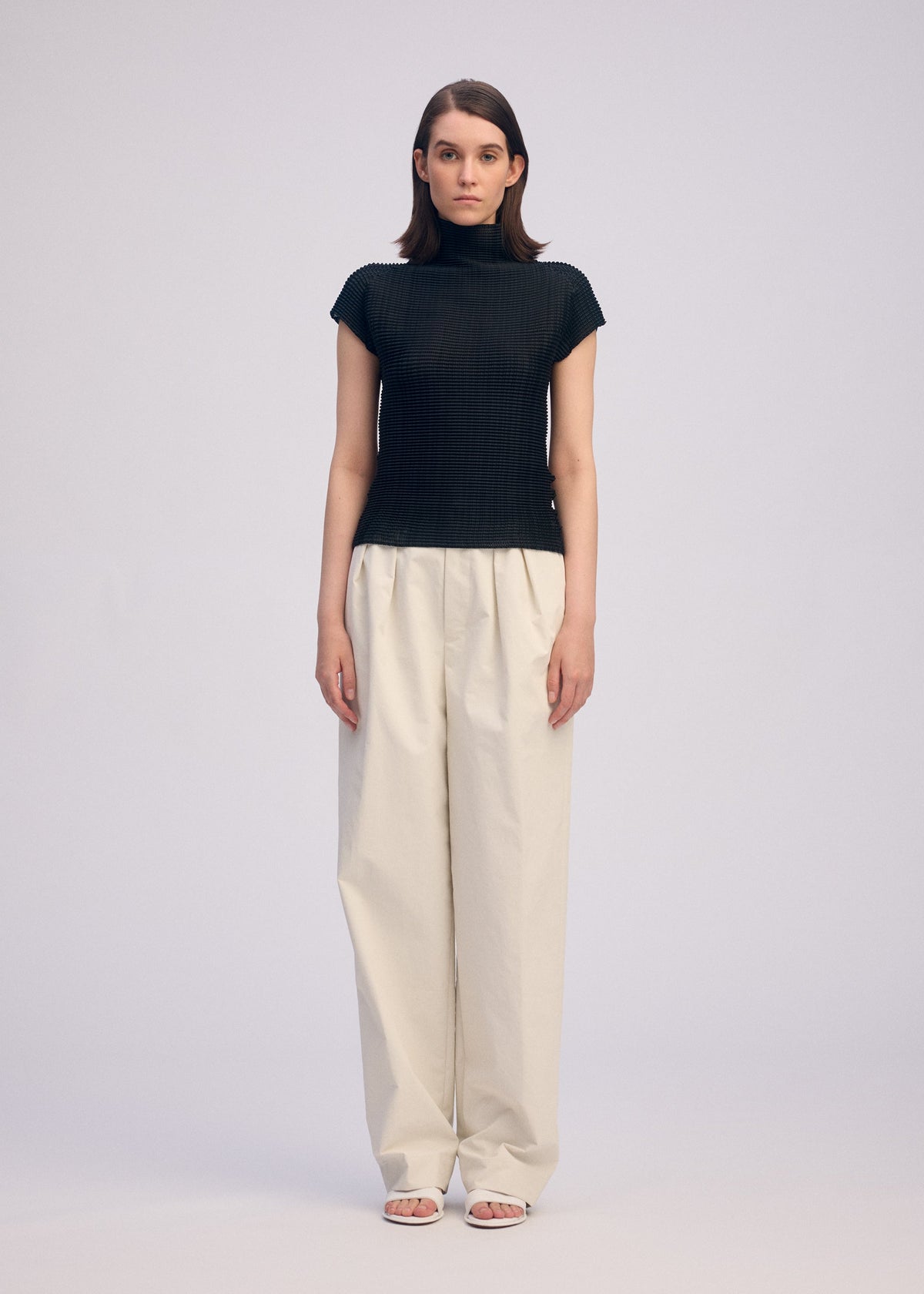 A woman faces forward against a plain background, wearing the ISSEY MIYAKE WOOLY PLEATS BK/WT-66— a black high-neck top with white loose trousers and open-toe sandals. Her expression is neutral, arms relaxed at her sides. view 4 of 7