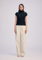 A woman faces forward against a plain background, wearing the ISSEY MIYAKE WOOLY PLEATS BK/WT-66— a black high-neck top with white loose trousers and open-toe sandals. Her expression is neutral, arms relaxed at her sides.