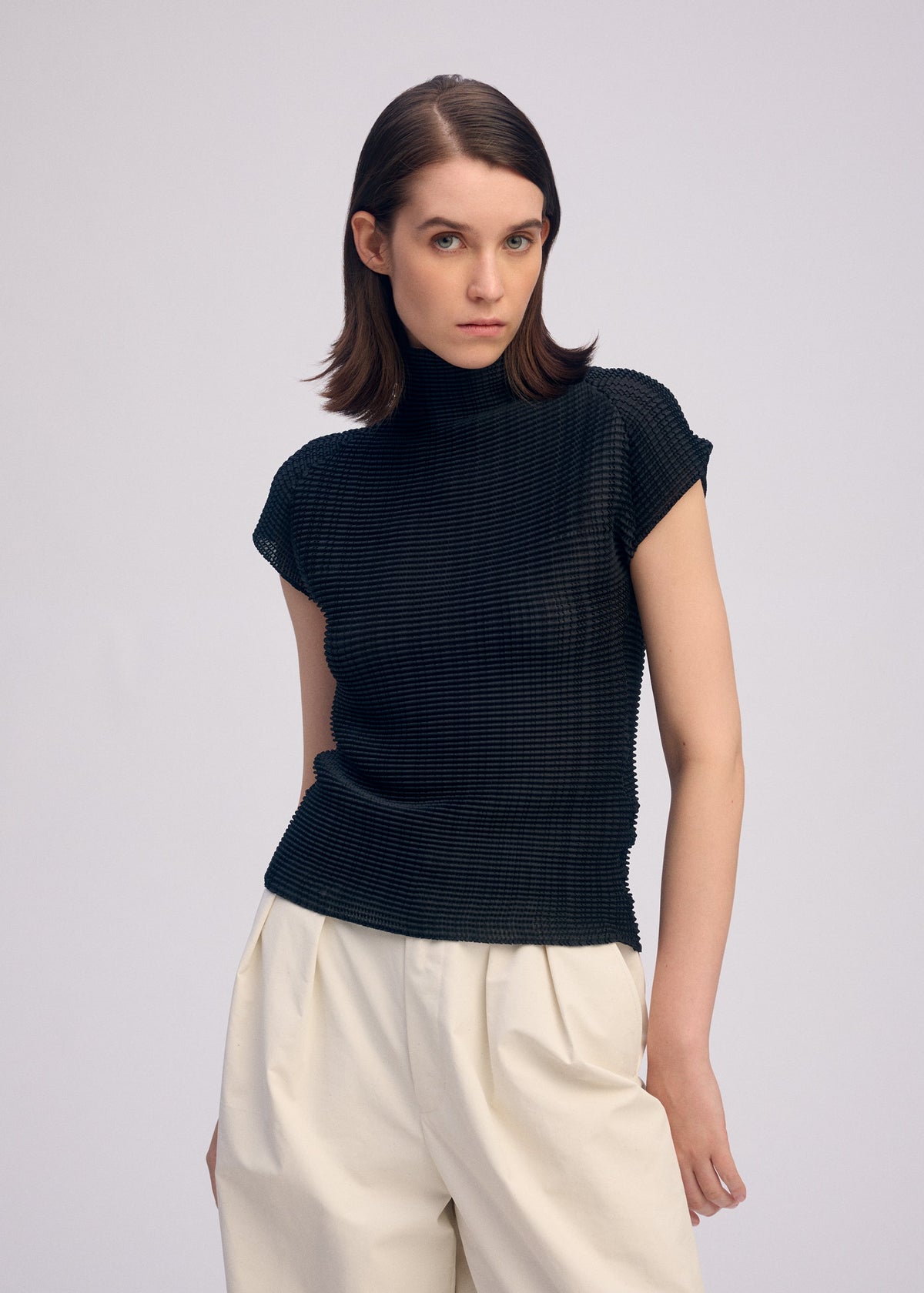 A woman with straight brown hair wears ISSEY MIYAKE's WOOLY PLEATS BK/WT-66, a stretchy high-neck top with French sleeves and loose, high-waisted cream trousers made from recycled polyester, standing against a plain light background. view 5 of 7