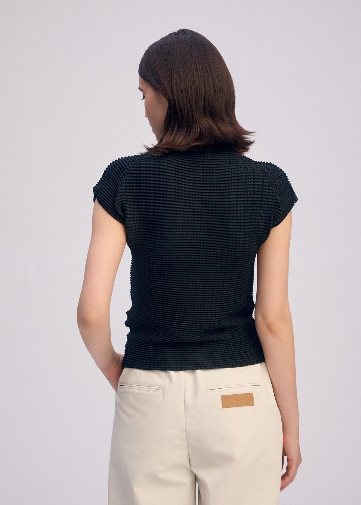 A person with short brown hair stands with their back to the camera, wearing ISSEY MIYAKE’s WOOLY PLEATS BK/WT-66 textured black high-neck top and high-waisted beige pants featuring a tan patch on the back pocket. view 6 of 7