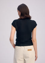 A person with short brown hair stands with their back to the camera, wearing ISSEY MIYAKE’s WOOLY PLEATS BK/WT-66 textured black high-neck top and high-waisted beige pants featuring a tan patch on the back pocket.
