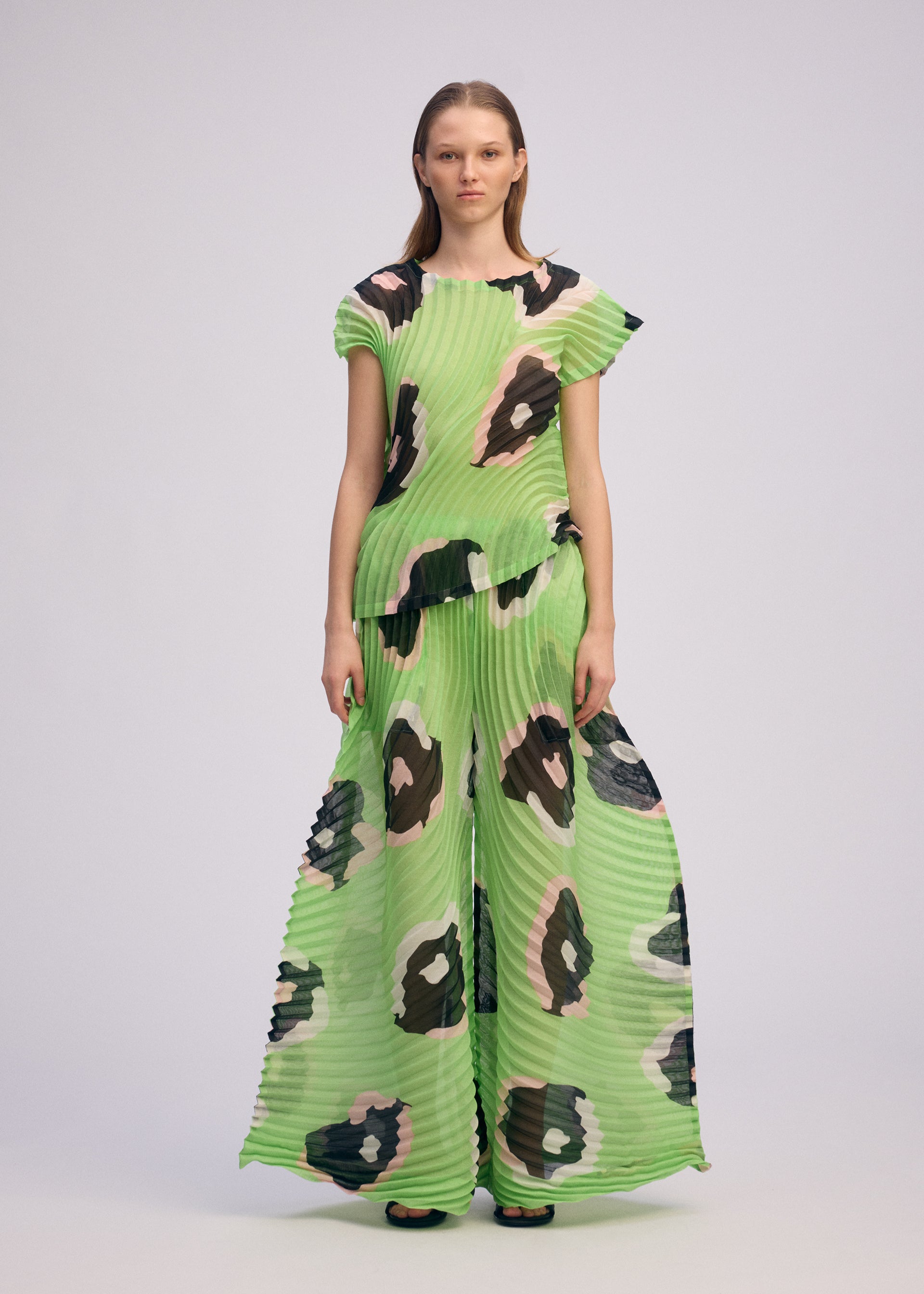 Zoomed view A woman stands against a plain background wearing ISSEY MIYAKE’s VERVE: a loose-fitting, pleated boat-neck top and wide-legged pants in lime green, featuring a bold black and brown abstract floral print.