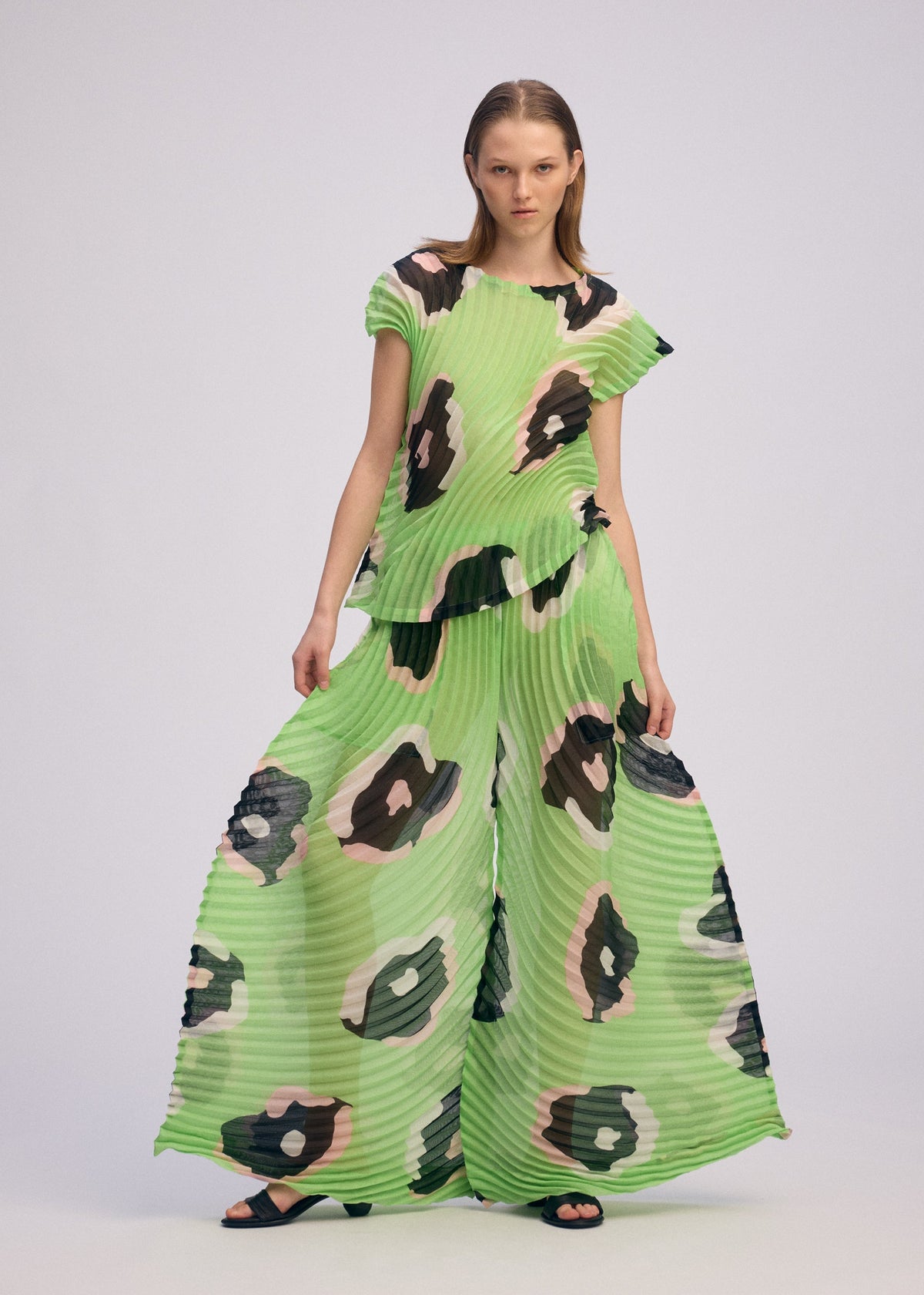 A woman stands confidently in VERVE by ISSEY MIYAKE, featuring a pleated boat-neck top and wide-leg pants in shimmering translucent green with bold abstract black, beige, and pink spots for a dramatic sculptural look. view 6 of 8