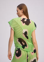 A person with straight brown hair wears the VERVE green pleated boat-neck top and shorts by ISSEY MIYAKE, featuring large abstract black, pink, and beige spots. They stand slightly turned away against a plain background.