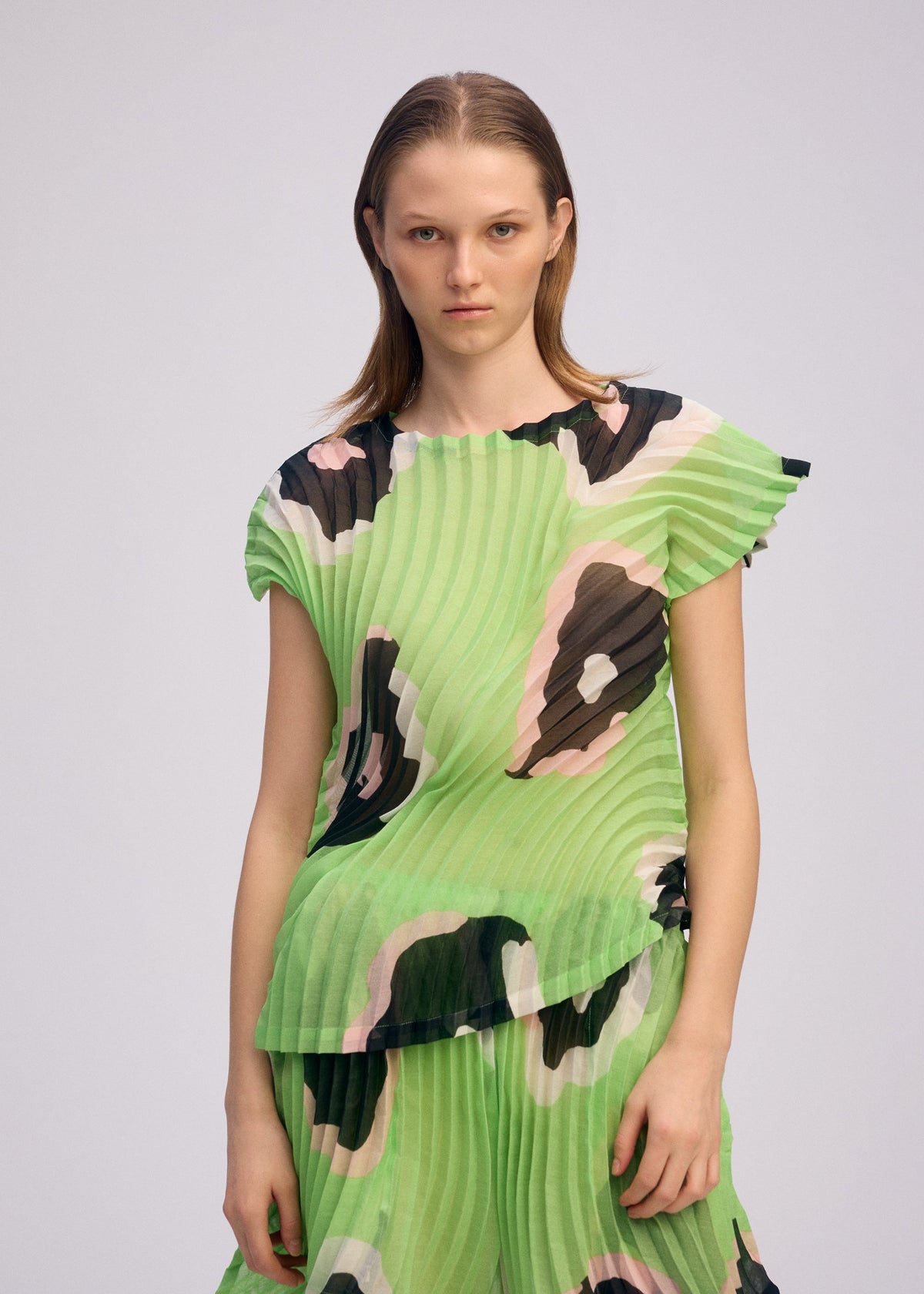 A person with straight, shoulder-length hair wears the VERVE pleated boat-neck top and matching skirt by ISSEY MIYAKE, featuring a wavy abstract black, pink, and white pattern, standing against a plain light background. view 8 of 8