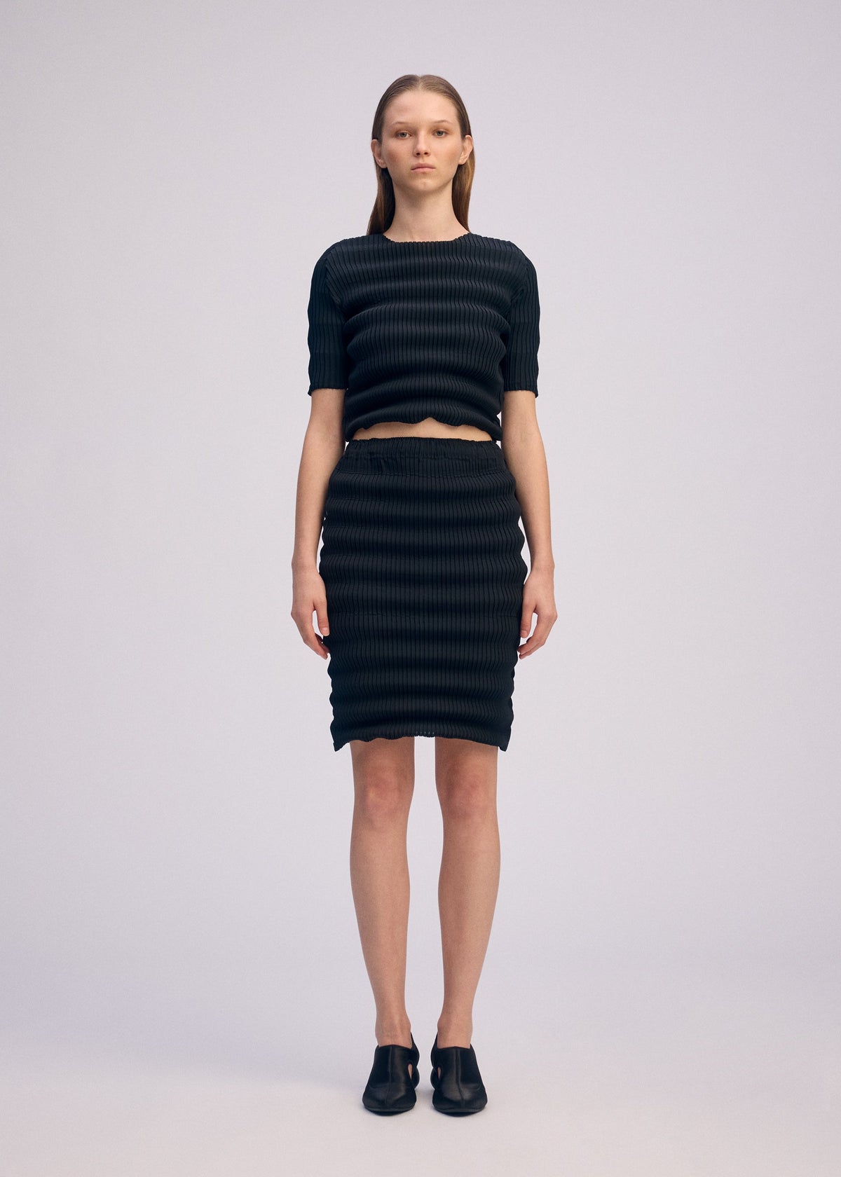 A person faces forward against a plain background, wearing ISSEY MIYAKE’s SPRING PLEATS: a black round-neck top and knee-length pleated skirt, paired with black shoes. Their straight hair is tucked behind their ears. view 4 of 6