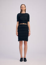A person faces forward against a plain background, wearing ISSEY MIYAKE’s SPRING PLEATS: a black round-neck top and knee-length pleated skirt, paired with black shoes. Their straight hair is tucked behind their ears.
