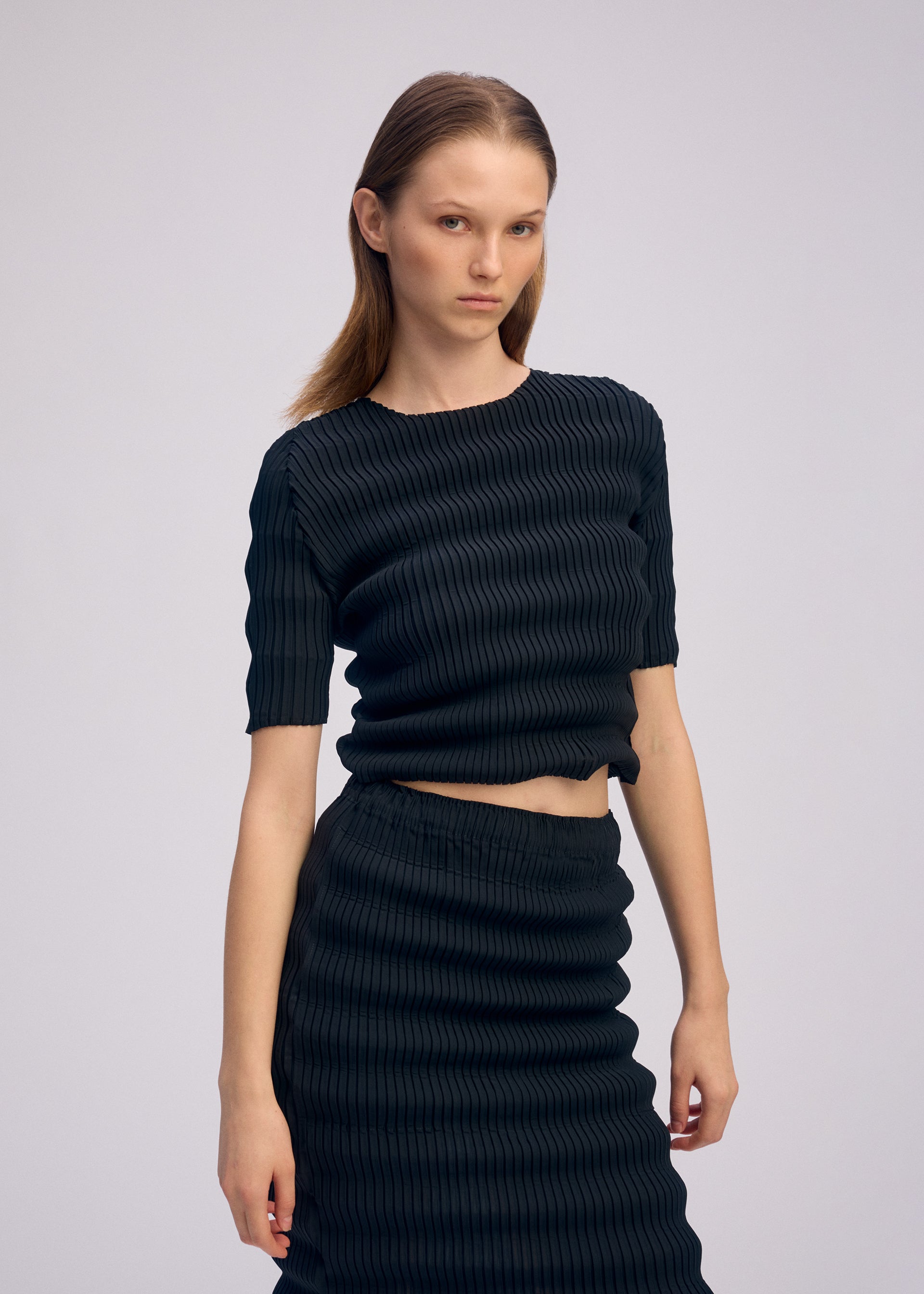 Zoomed view A woman with straight, light brown hair models the ISSEY MIYAKE SPRING PLEATS set—black ribbed-knit round-neck top and matching handmade-pleated skirt—against a plain background, looking serious with one hand by her side.