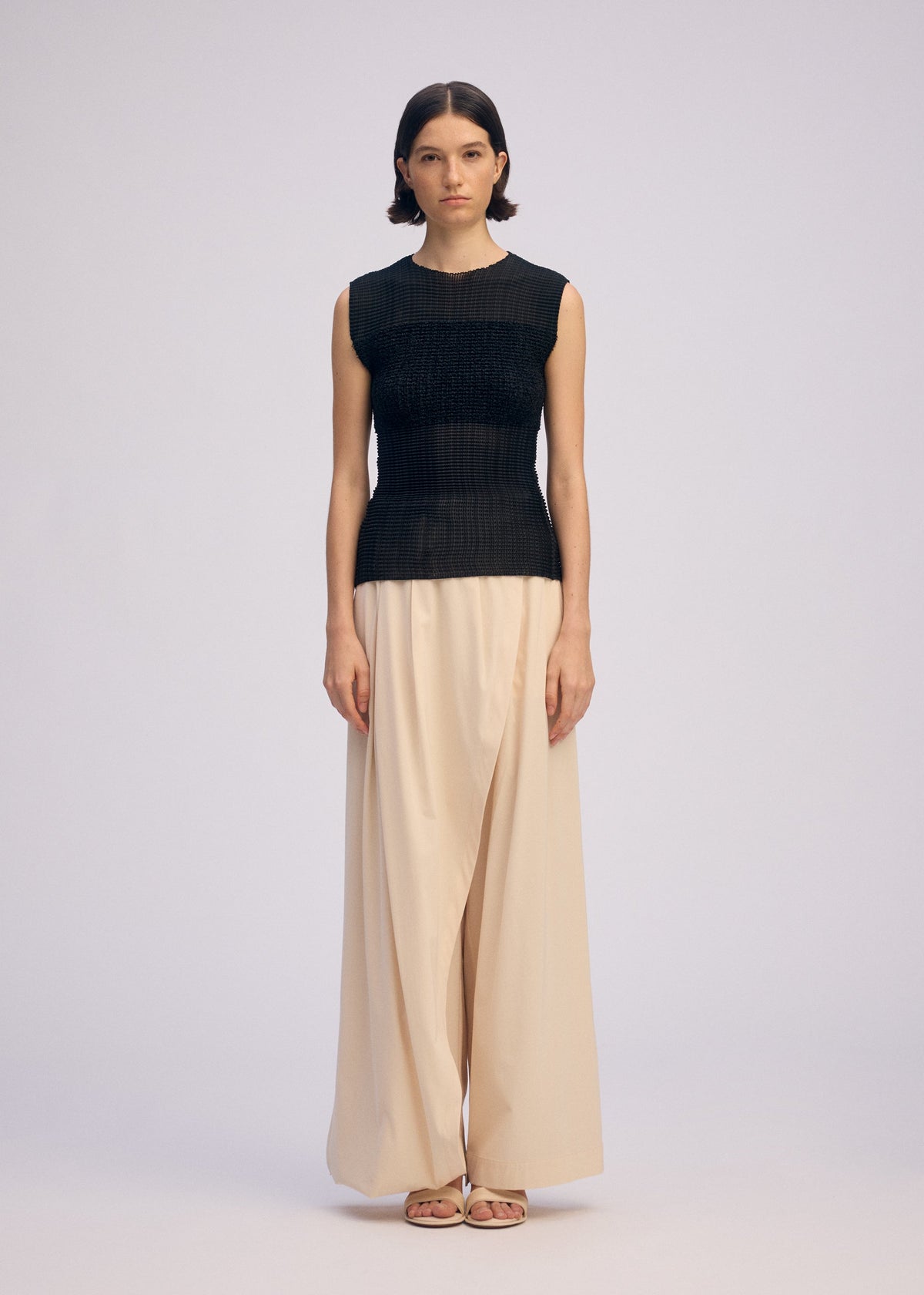 A woman faces forward in ISSEY MIYAKE's SHEER FUZZY PLEATS—a round-neck sleeveless black knit top and wide-legged cream trousers. She wears sandals, her straight dark hair behind her ears, with a neutral expression against a plain background. view 4 of 6