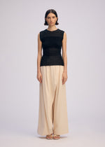 A woman faces forward in ISSEY MIYAKE's SHEER FUZZY PLEATS—a round-neck sleeveless black knit top and wide-legged cream trousers. She wears sandals, her straight dark hair behind her ears, with a neutral expression against a plain background.