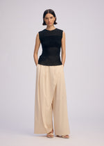A woman with short dark hair stands against a plain background wearing ISSEY MIYAKE’s SHEER FUZZY PLEATS: a black, round-neck sleeveless top in textured cut-pile, and cream high-waisted pleated wide-leg trousers, paired with sandals and hands in her pockets.