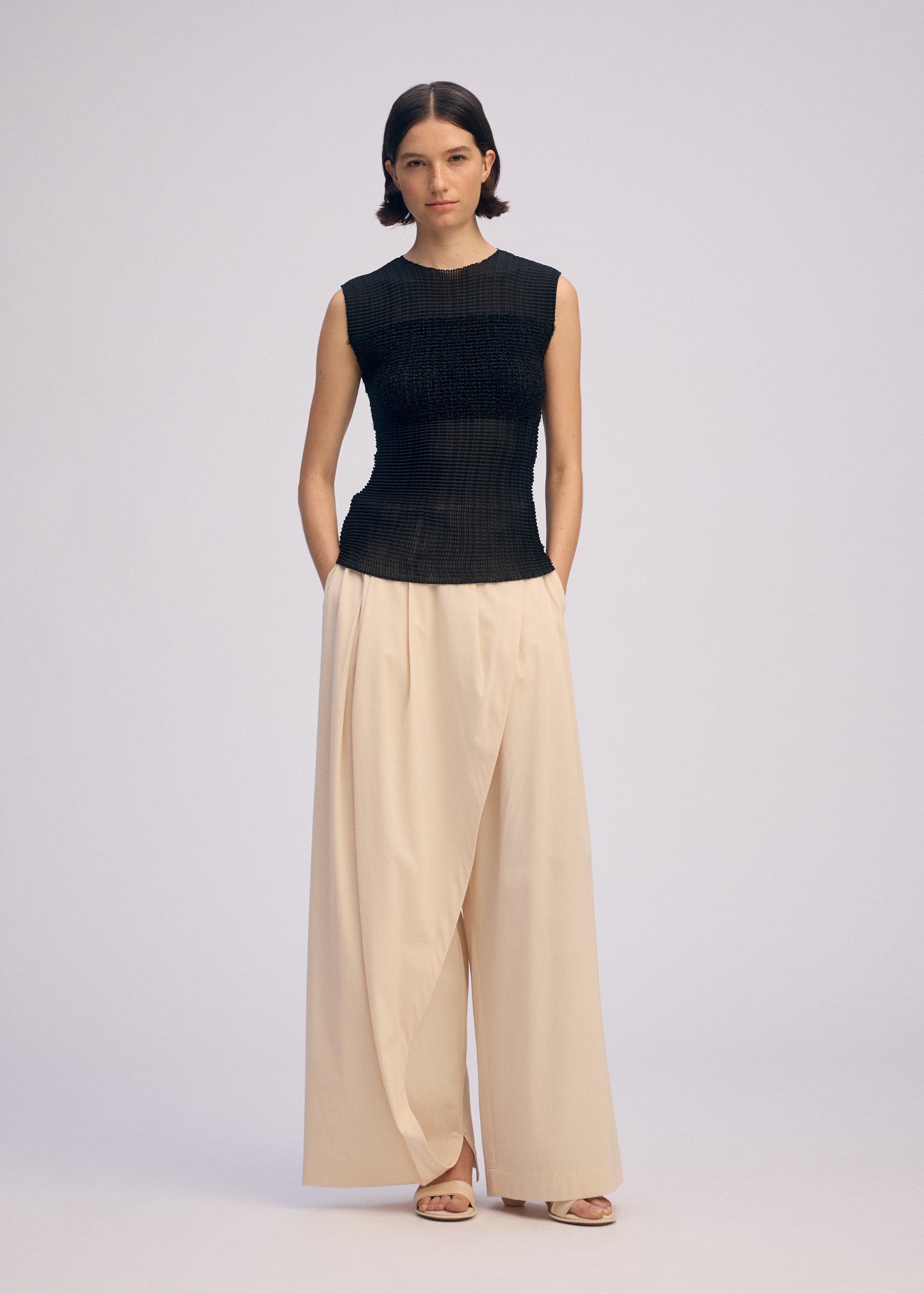 Zoomed view A woman with short dark hair stands against a plain background wearing ISSEY MIYAKE’s SHEER FUZZY PLEATS: a black, round-neck sleeveless top in textured cut-pile, and cream high-waisted pleated wide-leg trousers, paired with sandals and hands in her pockets.