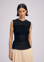 A woman with short brown hair models the ISSEY MIYAKE SHEER FUZZY PLEATS: a textured black sleeveless top and a light beige pleated skirt, standing against a plain background and looking directly at the camera with a neutral expression.