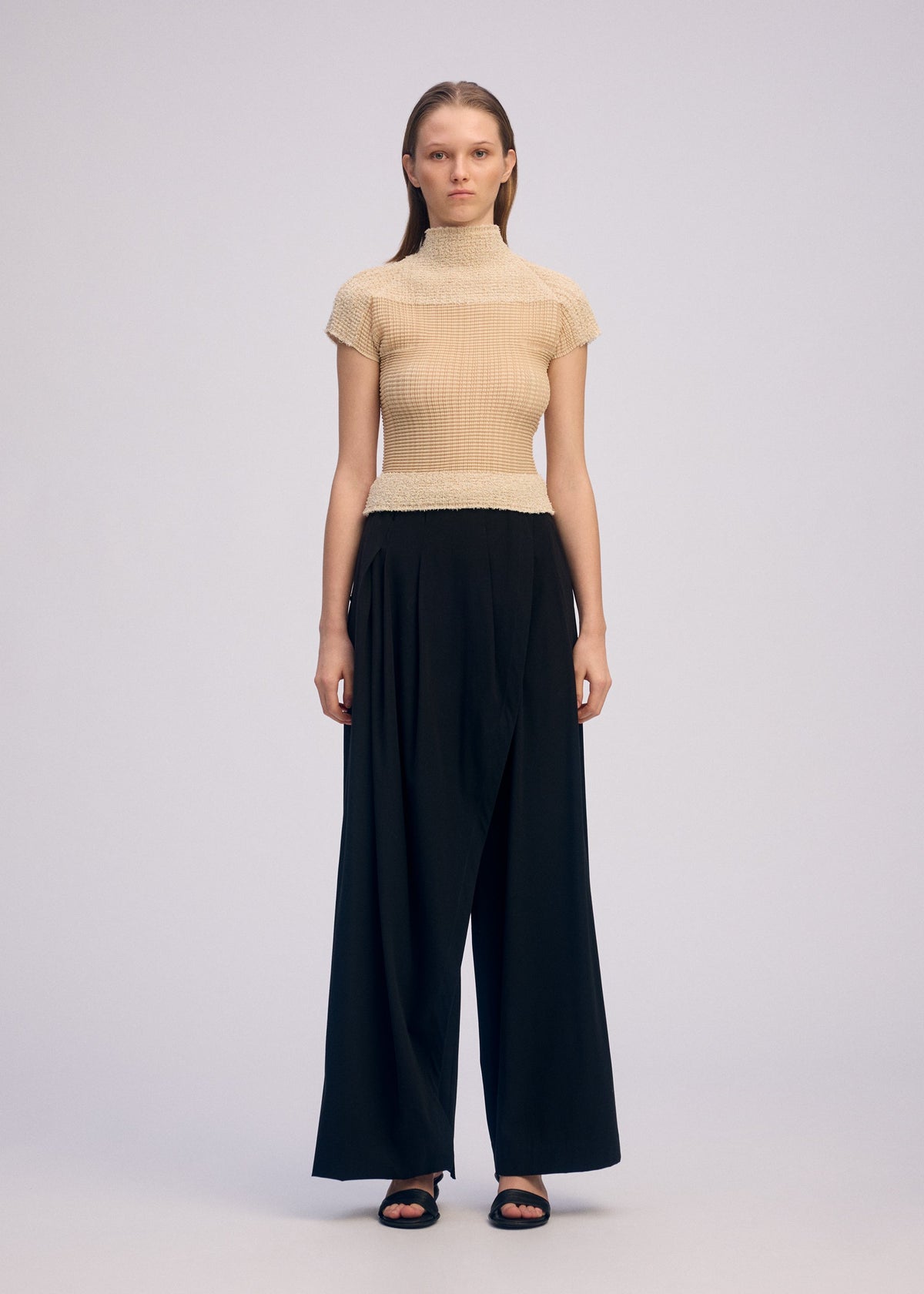 A woman stands facing forward against a plain light background, wearing ISSEY MIYAKE's SHEER FUZZY PLEATS: a beige high-neck top and loose-fitting black wide-leg pants paired with black sandals. view 4 of 7
