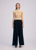 A woman stands facing forward against a plain light background, wearing ISSEY MIYAKE's SHEER FUZZY PLEATS: a beige high-neck top and loose-fitting black wide-leg pants paired with black sandals.