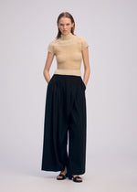 A woman with a neutral expression stands against a light background wearing ISSEY MIYAKE's SHEER FUZZY PLEATS: a beige French-sleeved high-neck top, black wide-leg high-waisted pants, and black open-toe sandals, hands in pockets.