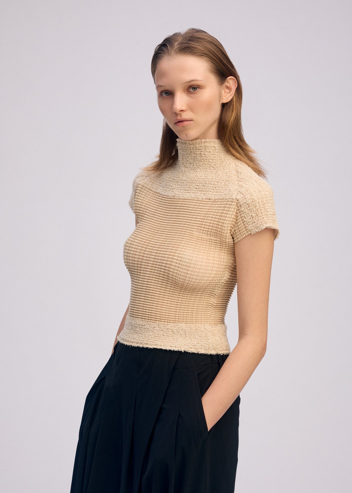A young woman stands with her hands in her pockets, wearing ISSEY MIYAKE's SHEER FUZZY PLEATS high-neck beige top and high-waisted black pants against a plain light background. view 6 of 7