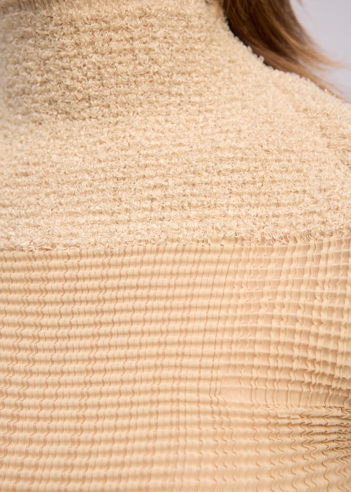 Close-up of ISSEY MIYAKE's SHEER FUZZY PLEATS: a beige, pleated cut-pile fabric with a grid-like texture, draped over the shoulder and neck as a French-sleeved high-neck top. Light brown hair partially visible. view 7 of 7