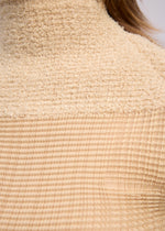 Close-up of ISSEY MIYAKE's SHEER FUZZY PLEATS: a beige, pleated cut-pile fabric with a grid-like texture, draped over the shoulder and neck as a French-sleeved high-neck top. Light brown hair partially visible.